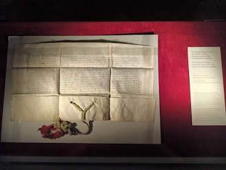 A historical document is displayed on a flat surface, consisting of aged, creased paper with handwritten script. Below the document, a decorative seal with green and red yarn is attached. To the right, a printed description on a board provides detailed context about the document, which appears to relate to a privilege issued by a king in 1433.