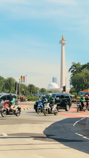A busy urban intersection with several motorcyclists wearing helmets and green jackets riding on the road alongside a black car. A tall monument with a golden flame at the top is prominently visible in the background, surrounded by lush green trees and clear blue skies.