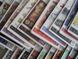 An array of colorful magazines or newspapers is spread out. Each publication features vibrant and varied cover designs, with a mix of illustrations, text, and patterns. The arrangement creates a visually dynamic and layered appearance.