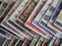 An array of colorful magazines or newspapers is spread out. Each publication features vibrant and varied cover designs, with a mix of illustrations, text, and patterns. The arrangement creates a visually dynamic and layered appearance.