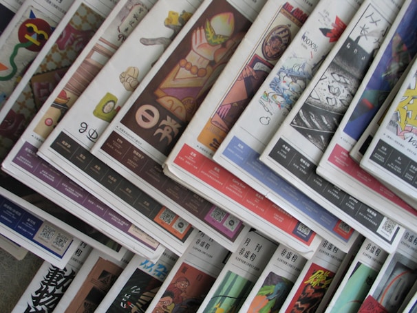 An array of colorful magazines or newspapers is spread out. Each publication features vibrant and varied cover designs, with a mix of illustrations, text, and patterns. The arrangement creates a visually dynamic and layered appearance.