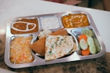 A stainless steel platter with six compartments filled with various Indian dishes. The food appears appetizing with different textures and colors. There are servings of dal with cream, yogurt or raita, curry, and biryani rice. The central compartment contains naan bread and a samosa. Another section has cucumber and carrot slices.