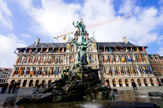 A large, ornate building with numerous international flags hanging from its facade stands behind a prominent fountain featuring a detailed statue. The sky is partly cloudy and the scene includes several people walking around the public square.