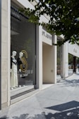 A storefront display featuring mannequins dressed in elegant, monochromatic outfits. The store window showcases the brand 'ST. JOHN' and is set against a backdrop of modern architecture with large glass panels. Trees cast shadows on the sidewalk, adding a natural element to the urban setting.