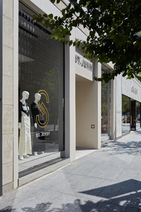 A storefront display featuring mannequins dressed in elegant, monochromatic outfits. The store window showcases the brand 'ST. JOHN' and is set against a backdrop of modern architecture with large glass panels. Trees cast shadows on the sidewalk, adding a natural element to the urban setting.