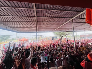 A large crowd of people is gathered under a metal-roofed shelter. Many individuals are raising their hands enthusiastically, and the crowd extends to an outdoor area with orange tents and trees in the background.