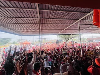 A large crowd of people is gathered under a metal-roofed shelter. Many individuals are raising their hands enthusiastically, and the crowd extends to an outdoor area with orange tents and trees in the background.