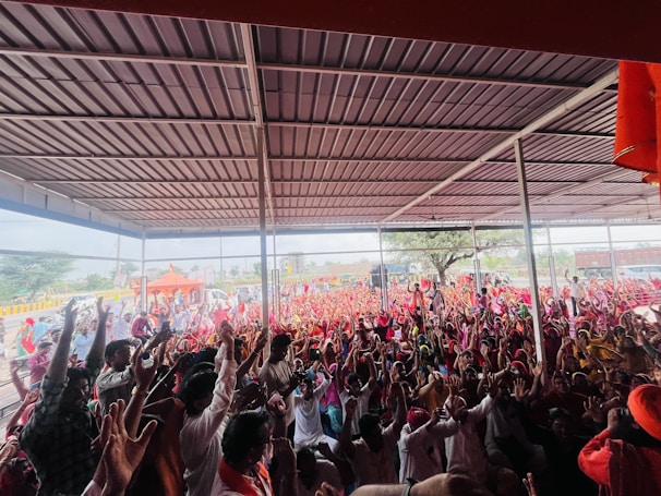 A large crowd of people is gathered under a metal-roofed shelter. Many individuals are raising their hands enthusiastically, and the crowd extends to an outdoor area with orange tents and trees in the background.