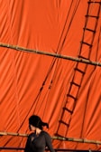 A person with dark hair is standing in front of a large, vibrant orange sail. The sail is made of fabric and is supported by wooden poles. Shadows of ropes create geometric patterns on the sail.