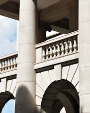 Detailed stone architectural elements with large columns, arches, and balustrades. The structure features a classic design with intricate details and shadows cast by the sunlight, highlighting the texture of the stone.