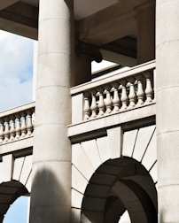 Detailed stone architectural elements with large columns, arches, and balustrades. The structure features a classic design with intricate details and shadows cast by the sunlight, highlighting the texture of the stone.
