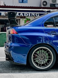 A close-up view of the rear side of a blue sports car featuring a large, black rear spoiler. The car's alloy wheel has a distinctive design, with multiple spokes and low-profile tires. Reflections on the car's glossy surface indicate a polished finish. In the background, there's a blurred sign with the word 'TRADING' partially visible.