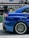 A close-up view of the rear side of a blue sports car featuring a large, black rear spoiler. The car's alloy wheel has a distinctive design, with multiple spokes and low-profile tires. Reflections on the car's glossy surface indicate a polished finish. In the background, there's a blurred sign with the word 'TRADING' partially visible.