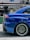 A close-up view of the rear side of a blue sports car featuring a large, black rear spoiler. The car's alloy wheel has a distinctive design, with multiple spokes and low-profile tires. Reflections on the car's glossy surface indicate a polished finish. In the background, there's a blurred sign with the word 'TRADING' partially visible.