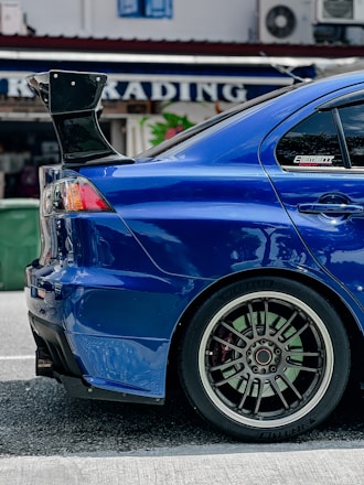 A close-up view of the rear side of a blue sports car featuring a large, black rear spoiler. The car's alloy wheel has a distinctive design, with multiple spokes and low-profile tires. Reflections on the car's glossy surface indicate a polished finish. In the background, there's a blurred sign with the word 'TRADING' partially visible.