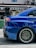A close-up view of the rear side of a blue sports car featuring a large, black rear spoiler. The car's alloy wheel has a distinctive design, with multiple spokes and low-profile tires. Reflections on the car's glossy surface indicate a polished finish. In the background, there's a blurred sign with the word 'TRADING' partially visible.