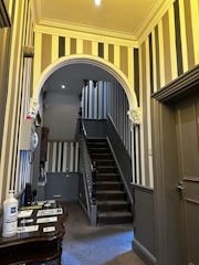 A hallway featuring bold vertical stripes on the walls, with a central staircase leading upwards. The staircase has dark carpeting and wooden banisters. The ceiling is adorned with decorative moldings, and there are wall sconces for lighting. On the left is a small table with hand sanitizer and informational brochures.