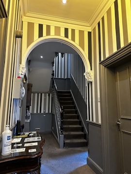 A hallway featuring bold vertical stripes on the walls, with a central staircase leading upwards. The staircase has dark carpeting and wooden banisters. The ceiling is adorned with decorative moldings, and there are wall sconces for lighting. On the left is a small table with hand sanitizer and informational brochures.