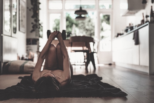 A person is lying on their back on a mat in a cozy, light-filled room. The room features a kitchen area and dining table with chairs. Soft lighting from large windows creates a calm atmosphere.