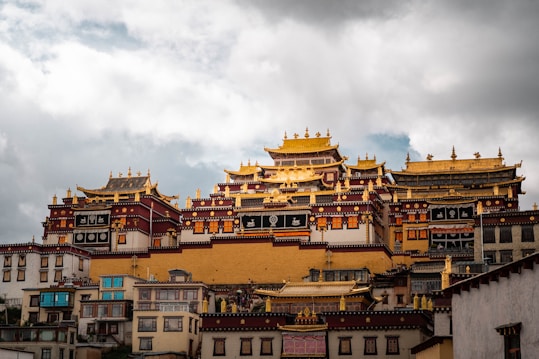 A large, ornate Tibetan-style monastery with multi-tiered roofs and intricate golden decorations stands majestically against a backdrop of a cloudy sky. The structure is a blend of white, red, and gold, and it includes various windows and banners with symbolic designs.