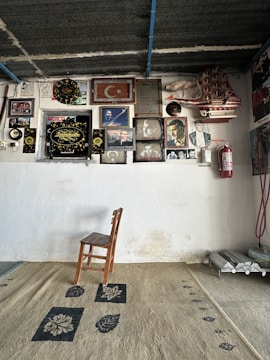 A small wooden chair sits on a patterned rug placed against a white wall. The wall is covered with various framed photographs, plaques, a model ship, a fire extinguisher, and the Turkish flag. Additionally, there is some electrical wiring visible on the right.