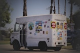 A white vehicle, resembling an ice cream truck, is decorated with various colorful posters and signs, including 'Stop for children' and 'Children Crossing'. The truck is parked on a road with palm trees in the background and some greenery at the sides.