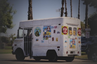 A white vehicle, resembling an ice cream truck, is decorated with various colorful posters and signs, including 'Stop for children' and 'Children Crossing'. The truck is parked on a road with palm trees in the background and some greenery at the sides.
