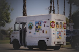 A white vehicle, resembling an ice cream truck, is decorated with various colorful posters and signs, including 'Stop for children' and 'Children Crossing'. The truck is parked on a road with palm trees in the background and some greenery at the sides.