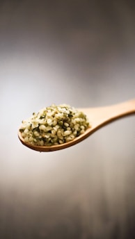 A wooden spoon filled with hemp seeds is suspended against a neutral, softly blurred background. The seeds have a pale green and beige hue and a small, round shape.