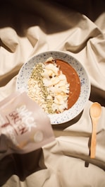 A bowl of smoothie topped with various ingredients like oats, coconut flakes, and seeds is placed on a patterned fabric. A wooden spoon lies next to the bowl, and a packet labeled 'Snack Garden' is partially visible on the side.