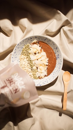 A bowl of smoothie topped with various ingredients like oats, coconut flakes, and seeds is placed on a patterned fabric. A wooden spoon lies next to the bowl, and a packet labeled 'Snack Garden' is partially visible on the side.