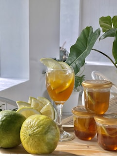 A refreshing glass of iced tea garnished with slices of lemon is set on a wooden surface. Next to the glass, there are two whole green lemons and a set of lemon wedges. Additionally, there are three jars filled with a tea-like liquid with lemon slices, accompanied by green leafy plant decorations in the background.