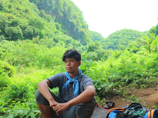 A person sits among lush green foliage, appearing relaxed and contemplative. They are wearing a gray shirt and a blue scarf around their neck. A colorful backpack is placed nearby, and the surrounding area is filled with dense trees and greenery, suggesting a natural, outdoor setting.