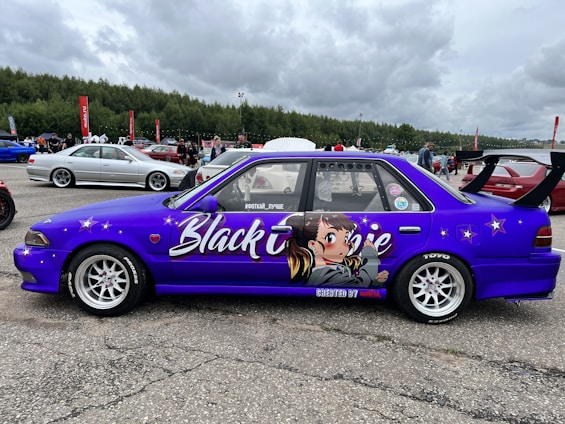 A modified purple car is prominently displayed, featuring distinctive anime-style artwork on its side. The design includes a large character illustration along with the words 'Black Lime'. The car has a large rear wing and custom wheels. Multiple other cars and people are visible in the background, suggesting a car show or event.