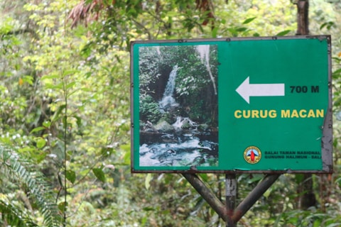 A signpost in a lush, green forest setting indicating the direction to a location called Curug Macan. The sign is predominantly green with white and yellow text, showing an image of a waterfall and a distance of 700 meters.