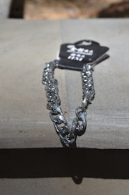 A metallic silver chain is placed on a flat surface under direct light. The chain is attached to a black tag with some text on it.