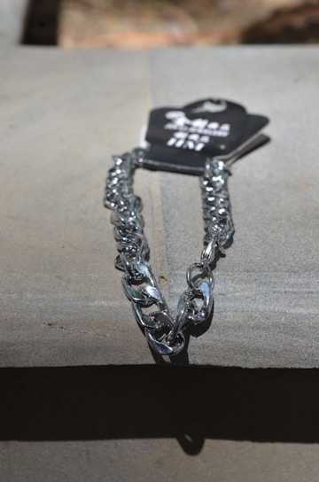 A metallic silver chain is placed on a flat surface under direct light. The chain is attached to a black tag with some text on it.