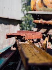 Rusty metal beams and industrial equipment are stacked in an outdoor setting. The beams are weathered and show signs of wear and tear, with a focus on a yellow beam in the foreground. The background suggests an industrial environment surrounded by greenery.