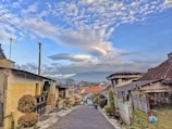 A narrow cobblestone street lined with houses on both sides, some with traditional tiled roofs. The sky is clear with a few scattered clouds and an unusual cloud formation that resembles a UFO. There are a few plants and small trees placed along the sides of the street. In the distance, there are more houses with orange rooftops and what appears to be mountains or hills in the background.