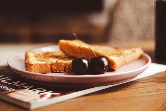 Two slices of toasted bread are placed on a light pink plate with two dark cherries on top. The plate rests on a wooden surface and sits on top of a magazine with partially visible text. The background appears softly blurred, giving a cozy and warm ambiance.