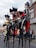 A group of performers dressed in elaborate, theatrical costumes involving large hats and face paint are playing musical instruments, notably drums and cymbals, while standing on stilts. They are performing outdoors in what seems to be a street, with onlookers in casual clothing watching them. The background includes buildings and store signage.