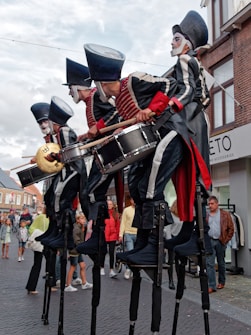 A group of performers dressed in elaborate, theatrical costumes involving large hats and face paint are playing musical instruments, notably drums and cymbals, while standing on stilts. They are performing outdoors in what seems to be a street, with onlookers in casual clothing watching them. The background includes buildings and store signage.