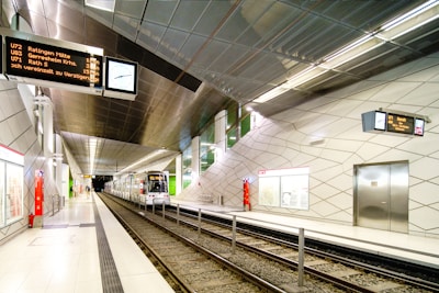 An underground train station with sleek modern design features. The platform is clean, with geometric patterns on the walls and a metal ceiling. A train is approaching on the track, and digital display signs show travel information. There are few people visible, adding to the calm and orderly atmosphere.