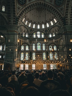 An ornate interior with a high, intricately decorated dome, stained glass windows, and numerous hanging lights. A large crowd of people is gathered, suggesting a communal or religious gathering.
