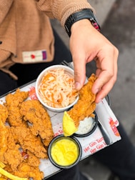 A hand holding a piece of fried chicken being dipped into a creamy yellow sauce. The meal is served on a tray containing more pieces of crispy fried chicken alongside small bowls of coleslaw and different sauces. The person is wearing a brown jacket with a visible pattern and a black watch.