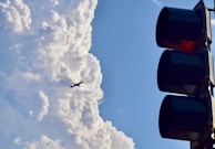 A silhouette of a commercial airplane flies against a backdrop of large, fluffy clouds. On the right side, a traffic light with red light illuminated stands out against the sky.