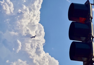 A silhouette of a commercial airplane flies against a backdrop of large, fluffy clouds. On the right side, a traffic light with red light illuminated stands out against the sky.