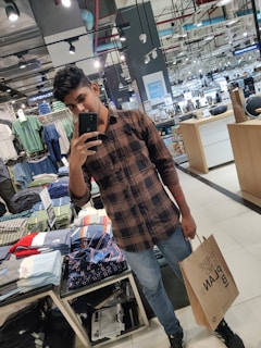 A person is taking a selfie in a clothing store surrounded by neatly arranged folded clothes. The individual is holding a brown shopping bag and wearing a brown checkered shirt and blue jeans. The store has bright lighting and several mirrors along the walls.