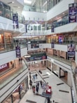 A multi-level shopping mall with large glass railings and visible store fronts including fashion retailers. People are walking throughout the space, with some carrying shopping bags. Sales banners are prominently displayed.