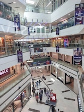A multi-level shopping mall with large glass railings and visible store fronts including fashion retailers. People are walking throughout the space, with some carrying shopping bags. Sales banners are prominently displayed.